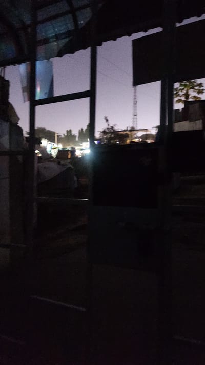The image appears to be taken at dusk or nighttime, as indicated by the dim lighting and visible streetlights and artificial illumination in the background. The scene is partially obscured by what seems to be a roof or overhang in the foreground, creating a framed view of the environment.

The main subjects visible are reflections in a window or glass surface, depicting a street scene with buildings and trees. A construction crane is visible in the distance, suggesting ongoing development or infrastructure work. In the lower portion of the image, there are lights from vehicles or buildings, adding to the urban or semi-urban setting. A white object, possibly a dog or a sheet, is partially visible in the middle ground, near some structures. The overall atmosphere is quiet and possibly a bit hazy due to the lighting conditions.

The location context provided is Kipala Mpakani, Tanzania, which helps to contextualize the imagery as likely depicting a scene from that region. There is no visible text in the image.