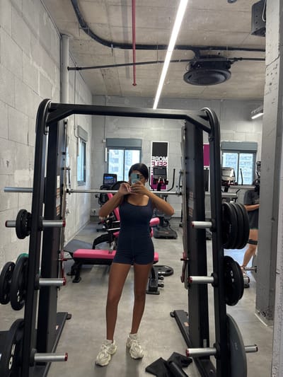 The image shows a woman standing in a gym in Ramla, Israel, located at latitude 31.931 and longitude 34.85. She is wearing a dark blue one-piece workout outfit and white sneakers. She is holding a smartphone in her right hand and taking a selfie. There is a weight rack behind her and a pink bench in the background. There are several other pieces of gym equipment visible in the image, including treadmills and elliptical trainers. There is a mirror on the right side of the image reflecting the woman and some of the gym equipment. In the reflection, a person is visible in the distance, partially obscured, wearing a gray shirt. The gym has white tiled floors and exposed concrete ceilings with fluorescent lights. The room is spacious and well-lit.