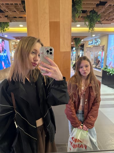 Two females are reflected in a mirror. The taller female holds a smartphone, taking a selfie with the other female in the reflection. The mirror reflects a store within a mall setting. The location of the photo is unknown.