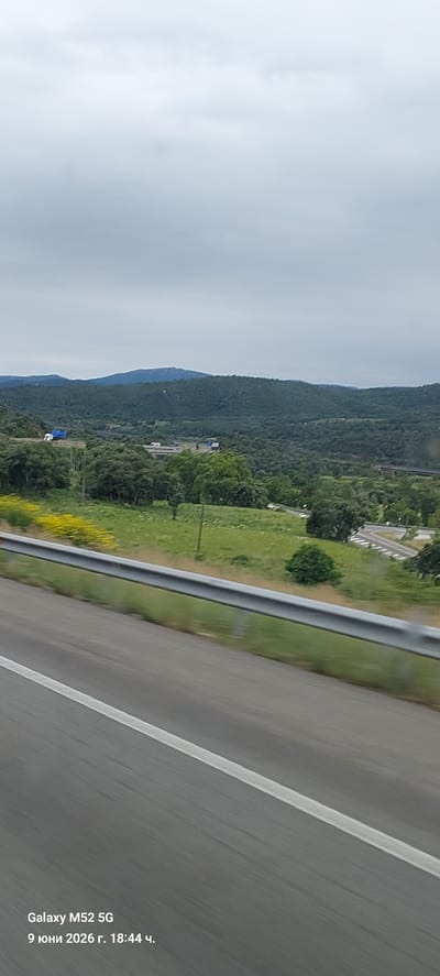 The image depicts a panoramic view of a rural landscape with rolling hills covered in dense green foliage under an overcast sky. In the foreground, a paved road with a white dividing line and a silver guardrail runs diagonally from the bottom left to the top right of the frame. Beyond the road, a grassy field is visible, punctuated by scattered trees and a patch of vibrant yellow wildflowers. Further in the distance, there is evidence of infrastructure, including what appears to be a highway with vehicles, and a bridge or overpass. The overall scene suggests a scenic drive through a natural environment. The bottom left corner of the image contains a timestamp and device information: "Galaxy M52 5G 9 юни 2026 г. 18:44 ч." which translates to "Galaxy M52 5G June 9, 2026 18:44". This information, combined with the visual elements, places the scene in a natural, possibly mountainous or hilly area, likely on the outskirts or in the vicinity of La Jonquera, Spain, given the mountainous terrain and potential for highways connecting major regions.