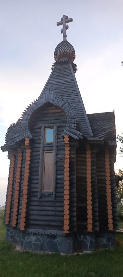 A dark wooden chapel with a cross on top is shown in this image. The chapel is made of logs and has a pitched roof with shingles. There is a small window on the front of the chapel. The chapel is surrounded by grass. The sky is a light blue. The image appears to have been taken during the day.