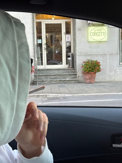 The scene is viewed from inside a vehicle, looking out a side window at a street in Pedaso, Italy. The prominent subject is a light gray stucco building with a white double-door entrance featuring glass panels. Above this entrance, a square, light green illuminated sign displays "dal 1950 CONCETTI." The building number "10" is affixed to the wall just to the right of the door. Three gray steps lead from the gray tiled sidewalk to the entrance. To the right of the building's entrance, a large terra cotta pot contains red flowering plants. A metallic cylindrical ash receptacle stands to the left of the doors, which also exhibit a 'No Smoking' sign on the glass. In the immediate foreground, to the left, a person's head and upper torso are partially visible, obscured by a light-colored hooded garment. A hand with visible fingers and natural nails, partially covered by the cuff of a matching light-colored long-sleeved garment, holds a lit cigarette with a visible incandescent tip, positioned outside the vehicle's window frame. The vehicle's dark gray interior door panel and black window frame are visible in the lower right of the image.