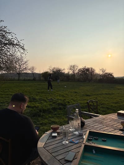An outdoor scene in Bû, France, depicts a golden hour setting with a low sun visible on the right horizon. In the foreground, a male with short dark hair, wearing a dark long-sleeved shirt, is seated with his back to the viewer at a weathered wooden octagonal patio table. He is looking down at a smartphone in his hands. The table holds a clear glass bottle, two empty patterned glasses, a small lighter-colored smartphone, an empty brown ceramic bowl, a small metal tin, a partially visible Marlboro Red cigarette pack, a small blue package, and an open green-lined backgammon board containing black checkers and two white dice. A wooden chair is pulled away from the table on the right.
In the midground, a large, well-maintained grassy lawn extends, where two individuals are present. One individual, positioned left of center, wears dark clothing and light shoes, appearing to be engaged with a white soccer ball situated on the grass in front of them. The second individual stands further back and to the right, wearing lighter clothing.
The background features a dense line of deciduous trees and bushes, some with emerging foliage, demarcating the end of the grassy area. A fence line is partially discernible behind the vegetation. The sky displays a gradient from light blue at the top to a warm orange-yellow near the horizon, where the circular bright yellow sun is prominent on the right.