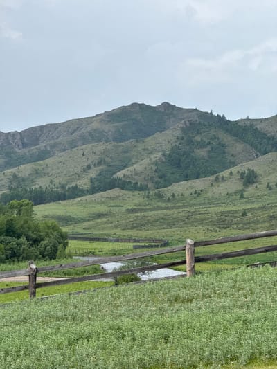 The image depicts a natural landscape under an overcast sky. In the foreground, a rustic wooden fence runs horizontally across the frame, constructed from rough-hewn logs. Beyond the fence, a body of water, likely a river or stream, meanders through a green meadow. Further in the distance, rolling hills covered in lush vegetation and scattered trees stretch towards a large mountain range in the background. The mountains are characterized by their rugged slopes, also dotted with trees. The overall scene suggests a rural or wilderness setting. The location is identified as Anchyl-Chon, Russia. There are no people or visible activity in the image.