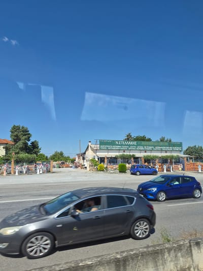 The image captures a sunlit scene on a road in Kardia, Greece, with a bright blue sky overhead. In the foreground, a dark gray hatchback car is driving on the right lane of the road. Its driver, a person with visible arms, is operating the vehicle. To the right, a bright blue hatchback car is also in motion on the same lane, with its driver partially visible. Further in the background, a smaller blue hatchback car is parked on the side of the road.

Adjacent to the road, there is a business with a long, green sign displaying text in Greek, "Ν. ΣΤΕΛΛΑΚΗΣ", and additional descriptive text below. This business appears to be a retail establishment selling ceramic items, with numerous terracotta pots and decorative pieces displayed outside. A red-tiled roof of a building is visible to the left of the business, and several tall green trees stand behind it. The overall environment suggests a commercial area located along a main road.