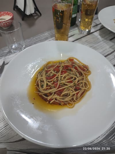 This image shows a plate of spaghetti with tomatoes and herbs, served in a light olive oil sauce. The pasta is presented in the center of a large white plate, which is placed on a striped placemat. Two glasses of beer are visible in the background, along with a small decorative bowl with a red and white pattern containing a white substance, possibly a dip or condiment. A napkin holder with white napkins is also in the background. The scene appears to be set at a table, suggesting a meal is being served.
The lighting suggests it is either late afternoon or evening, as there is a warm glow and shadows. There are no people visible in the image. The setting seems casual, possibly a restaurant or a home dining area.
The bottom right corner of the image shows a timestamp: "22/06/2026 21:30".