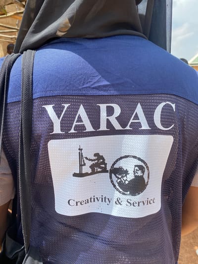 The image shows the back of a person wearing a dark blue vest with a white emblem. The emblem displays the text "YARAC" in large white letters at the top. Below this, there is a black and white graphic featuring two silhouettes: one of a person operating what appears to be a surveying instrument, and another of a person holding a camera. The phrase "Creativity & Service" is written in smaller white letters below the graphic. The vest has a mesh texture on the sides and is worn over a light-colored garment. The background is slightly out of focus but appears to be an outdoor setting with some natural elements like branches and what might be a building structure. The lighting suggests it is daytime. The person is wearing a black headscarf. The overall scene suggests the person is associated with an organization named YARAC and is possibly involved in some form of fieldwork or public engagement. The image does not contain enough information to determine the specific location in the city of Rim, Nigeria, nor does it provide details about specific actions or interactions beyond the visual representation on the vest.