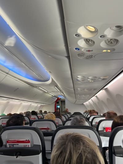 This image shows the interior of an airplane cabin, viewed from an angled perspective. The subjects are passengers seated in rows of seats, with their heads and upper bodies visible. The seats are modern and appear to be in a standard economy class configuration. The cabin walls are a clean white, with overhead compartments and ventilation systems visible. The lighting in the cabin is a soft white, with some areas illuminated by a blue glow, possibly from mood lighting.
The image depicts a scene of passengers seated in preparation for or during a flight. The atmosphere appears calm and ordinary, with no strong emotions readily apparent. There are some notable details: a "No Smoking" sign and a small circular fan are visible on the overhead panel. Several safety instruction cards are tucked into the seat pockets. Text is visible on some of these cards, including "norwegian.net" and "Safety Instructions". The time of day is not explicitly indicated, but the interior lighting suggests it could be evening or a dimly lit cabin. The location is Stockholm-Arlanda, Sweden, though this context isn't directly evident from the visual elements alone.