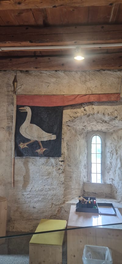 The image depicts an interior scene within a stone building, possibly a historic structure in Sargans, Switzerland. The dominant features are a large, rustic banner hanging on the textured stone wall, depicting a white bird with yellow feet against a dark background, bordered by a red fabric strip. To the right, a tall, arched window with a grid of panes lets in natural light. Below the window, on a wooden table, is an organizer filled with pens and pencils, a white paper, and a small green object. A wooden desk or cabinet is visible in the foreground. The ceiling is constructed of rough wooden beams. The overall impression is of an old, possibly fortified, interior space.
