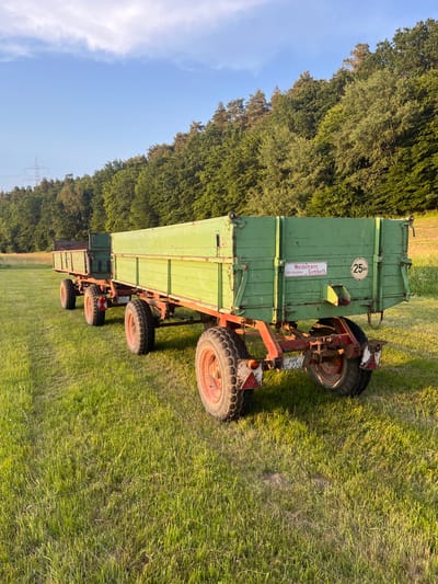 In Wiera, Germany, two green agricultural trailers are parked side-by-side in a grassy field. The trailers are attached to each other and are made of wood with red metal frames and wheels. The larger trailer in the foreground has a "25km" speed limit sticker and a license plate that reads "HR XC 220". The background features a dense forest and a clear blue sky with scattered clouds. The sun is setting, casting a warm glow on the scene. There are no people present in the image.