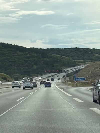 The image shows a view from a vehicle driving on a multi-lane highway in Randersacker, Germany. The highway is lined with trees on the left and a rocky embankment on the right. There are several cars on the highway, and a sign indicating "Ausfahrt" (exit) is visible on the right side of the road. The sky is cloudy.
