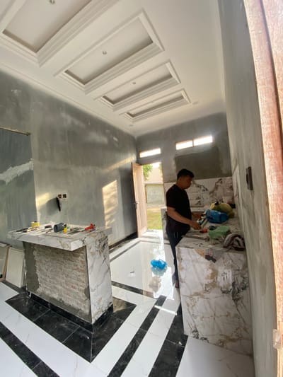 In an unfinished kitchen in Meunasahlhok, Indonesia, a man stands at a marble countertop, engaged in what appears to be home renovation or construction. The kitchen is in a raw state, with gray cement walls and a decorative, coffered ceiling. The floor features a bold black and white striped tile pattern. The man, wearing a black t-shirt and dark pants, is focused on the countertop, where various tools and materials are scattered, including a red-handled tool, a blue plastic bag, and what appears to be fabric. A partially constructed brick island is visible on the left. Sunlight streams through a doorway on the right, illuminating the scene and casting shadows. The surrounding environment suggests a residential construction site, with glimpses of the outdoors through the doorway.