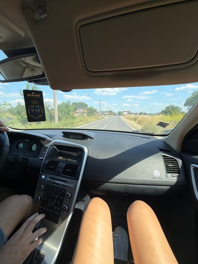 The image shows the interior of a car from the perspective of a passenger. The road ahead is visible, with greenery and houses on either side. The dashboard and steering wheel are in the foreground. There are two people in the car, one driver whose hand is on the gear stick and another passenger whose legs are visible in the lower part of the image. A car air freshener hangs from the rearview mirror. The scene appears to be in a rural area outside of Manastir, Bulgaria.