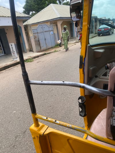 An outdoor, eye-level, medium shot captures a scene from inside a yellow auto-rickshaw, looking forward. The auto-rickshaw has a weathered exterior, with visible metal structures and some paint chips. In the foreground, a black metal bar extends horizontally across the frame. A dark holder for a cup or bottle is attached to the yellow frame. To the right, part of a light pink seat is visible.

Beyond the auto-rickshaw, a street runs through a residential area in Jos, Nigeria. Buildings with corrugated metal roofs line the street. One building on the left has a sign that reads "M & G LAUNDRY." A large grey metal gate is visible on the right side of the street.

A woman in a green and white patterned traditional dress walks away from the camera on the sidewalk. Her head is covered by a matching headscarf. She carries a white bucket and a dark bag. Behind her, another person in a red top is partially visible. In the background, a dark-colored car is parked on the right side of the street. The sky is overcast, suggesting a cloudy day. A reflection in the auto-rickshaw's side mirror shows a person taking a selfie.