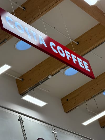 A tilted, upward-angled shot captures a red Costa Coffee sign suspended from a high ceiling. The white lettering of "COSTA COFFEE" is illuminated. The ceiling features a combination of bare wooden beams and smooth white panels with round windows. Linear lighting fixtures are also visible, casting bright light onto the scene. Part of a glass structure with metal supports is visible on the right side of the image. The location is suggested to be Pity Me, United Kingdom.