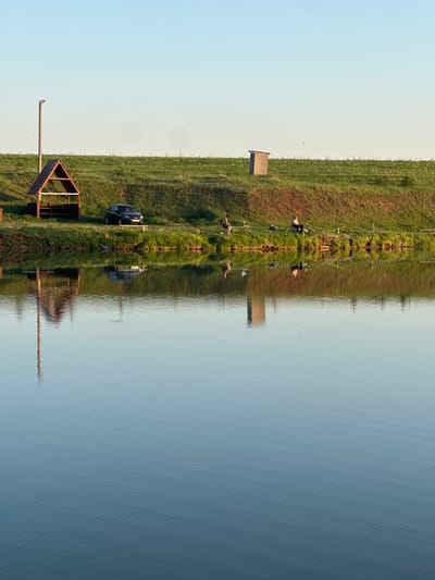 The image shows a landscape near Evlevo, Russia. A calm body of water reflects the scene, creating a mirrored effect. On the shore, there is a grassy area with a small wooden shed, metal structures (possibly fishing platforms), and a dark-colored car parked nearby. Several human figures, seemingly fishermen, are visible along the shoreline, some near fishing rods. The sky is light blue, suggesting either early morning or late afternoon light. The water's surface has subtle ripples, adding texture to the reflection.