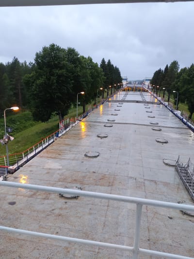 The image depicts a view from an elevated position, looking down a long, flat, wet, concrete surface that appears to be a lock chamber in a waterway or canal. The surface is marked with numerous circular hatches. On either side of the concrete expanse, there are railings with red and white accents. Along the edges, there are streetlights with orange lamps, some of which are reflecting on the wet surface. Beyond the railings and trees, a cloudy sky is visible. In the distance, there are structures that suggest a canal lock system, with yellow machinery and buildings. The surrounding environment consists of lush green trees and grass, indicating a natural setting. The overall scene is dimly lit, possibly due to overcast weather or it being twilight. The image does not contain any individuals or visible signs of activity. Based on the provided context, this scene is located in Alekseevskoe, Russia, and is part of the Vyshny Volochyok Water System, a canal lock on the Volga-Baltic Waterway.