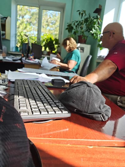 The image depicts an office scene with two individuals. In the foreground, a black baseball cap rests on a reddish-brown desk. To the left of the cap, a black keyboard is visible. Behind the keyboard, papers and other office supplies are spread across the desk. A computer monitor is partially visible on the left side of the frame. In the background, a woman in a teal shirt is seated, appearing to read or work on papers. She is positioned near a window. To the right of the woman, a bald man wearing glasses and a red shirt is visible from the side. He is seated and his arm is resting on the desk, near the cap. The environment appears to be an office with plants visible both inside and outside the window. The sunlight streams in through the window, creating shadows on the desk. The location is identified as Sovietskyi, Ukraine.