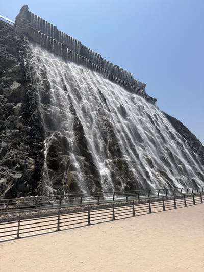 This is a low-angle, outdoor photograph capturing the Khor Fakkan Waterfall in Khor Fakkan, United Arab Emirates. The central and dominant feature is a large, multi-tiered artificial waterfall cascading down a dark, rugged rock face, which appears to be part of a mountain or engineered structure. Water flows in numerous parallel streams, creating a wide, visually dense sheet of water and generating significant spray that is visible against the dark rock. The top section of the structure features distinct horizontal ridges from which the water initially descends. The rock face itself is dark grey to black, exhibiting varied textures and natural fractures.
In the foreground, a light-colored paved walkway extends across the lower portion of the image, running parallel to the base of the waterfall. The paving stones are rectangular, laid in an interlocking pattern. A dark grey metal railing separates the walkway from the base of the waterfall. This railing consists of several horizontal bars supported by vertical posts, and its shadows are cast distinctly onto the paved surface, indicating direct sunlight from the upper left or behind the viewer. Behind the railing, at the base of the waterfall, a channel or pool collects the falling water. The upper right portion of the image reveals a clear, bright blue sky devoid of clouds. No individuals or animals are present in the scene.
