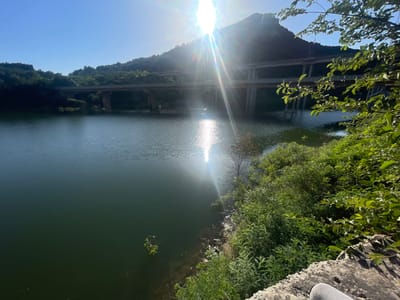 The image depicts a scenic view of the Asparuhovo dam and its surroundings. A clear blue sky dominates the upper portion of the frame, with the sun shining brightly, creating lens flares and a shimmering reflection on the water. In the background, a multi-tiered bridge spans across the dam, with lush green hills and trees forming a natural backdrop. The water of the dam is a deep, dark green, with a gentle ripple on its surface. In the foreground, a stone embankment is visible, with dense green foliage and bushes growing along the edge of the water. The overall scene suggests a tranquil and natural environment, likely a popular spot for outdoor recreation.