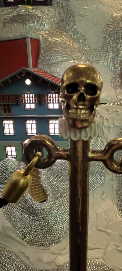 This is a close-up, eye-level, medium shot of a gold-colored decorative skull perched atop a stand with a ruffled collar. The skull is positioned in the foreground, its dark eye sockets and grin stark against the shiny metal. The stand is also gold, with decorative loops extending from it. In the background, a miniature blue and red house with white-trimmed windows is visible, nestled amongst a glittering, textured surface. The overall scene appears to be an artistic display, possibly within a shop or museum, given the detailed miniature architecture and the elaborate decorative elements. The location is confirmed to be the Swarovski Crystal Worlds in Wattens, Austria, a popular tourist attraction known for its art installations and crystal creations.