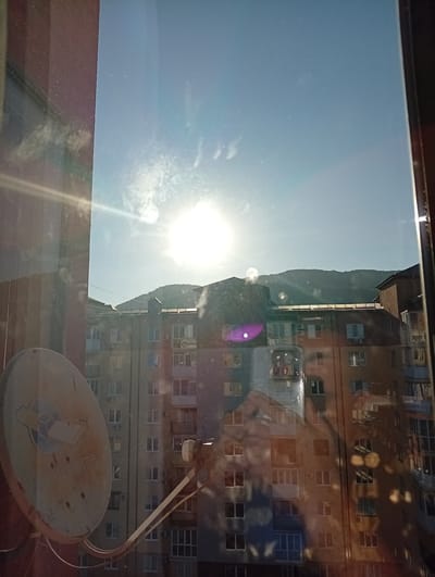 The image shows a view from a window in Agoy, Russia, overlooking a residential building and a mountain range in the background. The sun is bright and high in the sky, casting lens flares and reflections on the windowpane. In the foreground, a satellite dish is visible on the left side. The building appears to be several stories tall, with numerous windows and balconies. The sky is clear and blue. The overall scene depicts a sunny day in a somewhat urban or suburban environment.