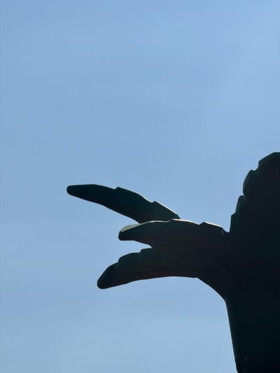A dark silhouette of a sculpture, possibly representing a bird's wing or a similar organic form, is positioned against a clear, pale blue sky. The sculpture is angled from the bottom right corner towards the left side of the frame, with two distinct curved sections visible in the foreground, resembling fingers or feathers. The overall impression is abstract and minimalist, emphasizing form and shadow. The location is Köprübaşı, Turkey.