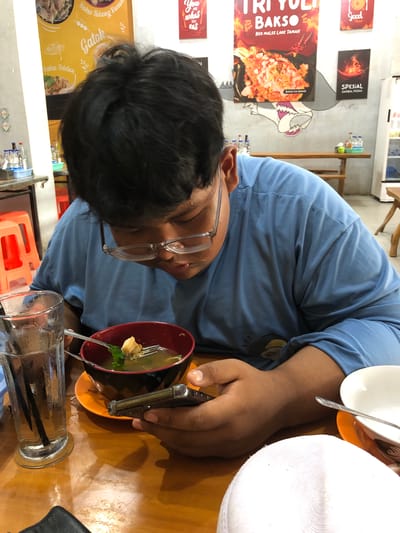 In a casual dining setting, likely a restaurant or food stall in Uteungeulinggang, Indonesia, a young person with dark hair and glasses is engrossed in their mobile phone while holding a fork over a bowl of soup. The person is wearing a blue long-sleeved shirt. The table is made of wood and holds a glass of water with a straw, the bowl of soup with a fork, and a white bowl. In the background, posters advertising food items like "Tri Yuli Bakso" and "Spesial Sambal Pedas" are visible on the wall, along with orange plastic stools and a refrigerator. The lighting suggests an indoor environment, possibly during the day or evening.