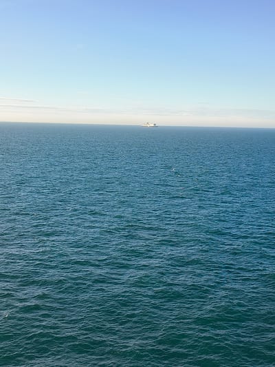 An image taken from a high vantage point shows a wide expanse of the sea under a clear blue sky with a thin band of clouds on the horizon. The water is a deep blue-green, with small ripples and waves visible across the surface. In the distance, a large white ship is visible on the horizon, appearing as a small speck against the vastness of the sea and sky. There are no people or any other discernible objects in the scene, creating a sense of isolation and open space. The location is identified as Unknown, United Kingdom.