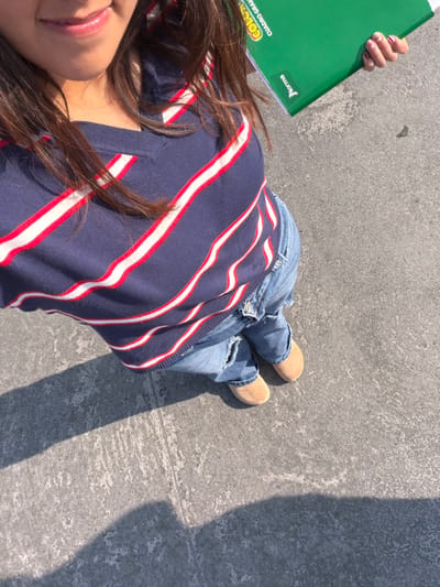 The image shows a person lying on a textured concrete surface, wearing ripped blue jeans and tan-colored shoes. They are wearing a navy blue top with horizontal red and white stripes. A green notebook is held up in their left hand. A shadow stretches across the concrete beside them. Their hair is dark brown. A portion of their face, including lips, is visible.