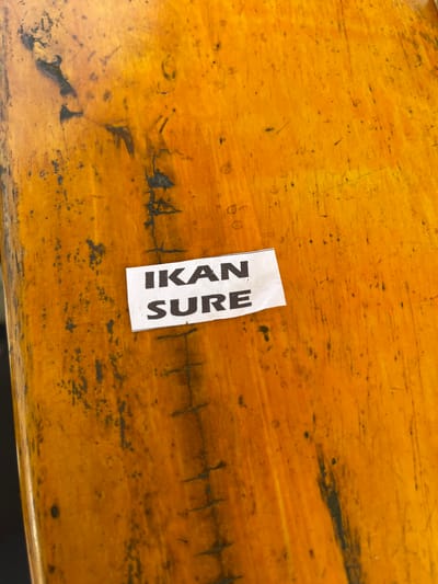A white paper label with black text is affixed to an orange wooden surface. The text on the label reads "IKAN SURE". The wooden surface exhibits a distressed finish with scratches and dark smudges. The image is a close-up, top-down view. The location is Julikeude Dua, Indonesia.