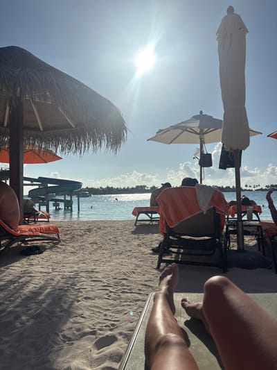 The scene depicts a sunny beach in Himmafushi, Maldives, viewed from a first-person perspective, looking out from a beach lounger towards the ocean. The foreground features the tanned legs of an individual resting on a dark-colored lounger, stretching towards the center of the frame.
The beach consists of light-colored sand with visible footprints and shadows. To the left, a large thatched beach umbrella provides shade, with part of an orange fabric umbrella visible behind it. On the right, a folded white beach umbrella stands upright, and further back, an open white beach umbrella is visible, from which a dark bag and a white bag hang.
Several individuals are present, mostly seated on beach loungers facing the water, their backs to the camera. One person on the far left is partially visible, while another on the far right has a leg raised. An orange towel is draped over a lounger in the mid-ground. A clear glass containing a light-colored beverage and a straw rests on a small side table next to a lounger on the right.
The mid-ground reveals calm, light blue ocean water shimmering with sunlight reflections. A multi-tiered, blue-grey water slide structure extends into the water from a platform on the left. A white boat is visible further out in the water, near the slide. At least one person is swimming in the ocean. The horizon features a distant landmass or island covered with green vegetation. The sky is clear blue with sparse white clouds on the horizon, and the bright sun, centered in the upper frame, produces a strong glare.