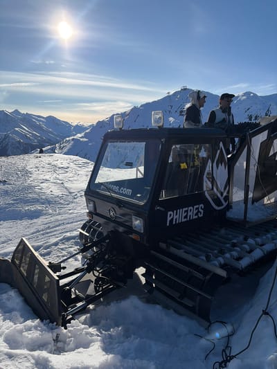 A black tracked snow groomer, prominently displaying "phieres.com" on its windshield and "PHIERES" with a stylized mountain logo on its side, is stationary on a snow-covered mountain slope in Schwendau, Austria. Its front plow is partially embedded in the snow. Two clear rectangular lights are mounted on the vehicle's cabin roof. On the elevated rear platform, two males are positioned behind DJ equipment. The individual on the left wears a light-colored hooded jacket and headphones, interacting with the equipment. The second male, positioned to the right, wears a dark cap, dark jacket, and headphones. Black and white patterned banners are affixed to the side of the platform. Black cables extend from the rear of the vehicle onto the snow. The scene is set against a clear, bright blue sky with a prominent sun in the upper left, illuminating extensive snow-capped mountain ranges in the background. A distant ski lift tower and cables are visible on a mountain peak.
