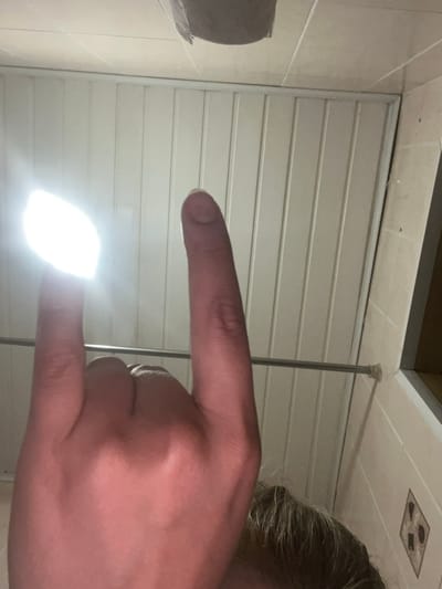 A close-up, low-angle shot captures a hand in the foreground, with its index finger pointing upwards and its thumb extended horizontally, forming a 'rock on' gesture. The index finger is illuminated by a bright light source, causing it to be overexposed and appear white. The background consists of a white, slatted ceiling and a horizontal metal rod, possibly a towel rack or shower rod, that spans across the frame. To the right, a portion of a tiled wall is visible. The image is not geo-located to Gorodets, Russia.