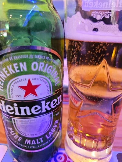A close-up image shows a Heineken beer bottle and a glass of beer positioned on a surface within an indoor setting in Audun-le-Tiche, France. The left subject is a green glass bottle of Heineken Original Pure Malt Lager, identifiable by its white and green label featuring the red star logo and "EST. 1873" text, along with "PURE MALT LAGER" and French descriptive text. The right subject is a clear glass beer mug, partially filled with a pale amber liquid topped by white foam and displaying active carbonation bubbles. The glass is embossed with a star pattern and has a "Heineken" logo faintly visible in reverse on its rim. The indoor environment features artificial, multi-colored lighting, predominantly blue, purple, and pink, which reflects on both the bottle and the glass. Beneath the items, a blurred surface displays red and white circular patterns, consistent with a coaster or table. No individuals are present.