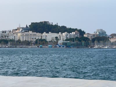 The image shows a cityscape on the coast of Cartagena, Spain. The foreground is dominated by the choppy, dark blue water of the harbor, with a light-colored concrete structure partially visible in the bottom left corner. Across the water, a dense line of buildings forms the city skyline.
To the left, there are several modern, multi-story apartment buildings with white facades and numerous windows. Behind them, on a hill, are more buildings, some with red-tiled roofs, and a prominent green expanse of trees. On the far left, a tall, open structure, possibly a crane or part of a construction, is visible.
In the center of the image, a large, white, neoclassical building with arched windows stands prominently. To its right, a series of white buildings of varying heights are clustered together, with some trees interspersed. Further to the right, a distinctive modern building with a faceted, glass-like facade and a stepped design stands out. Beyond this, more buildings and the natural contour of a hill are visible.
The background is a pale, hazy sky. Along the waterfront, a row of boats, primarily sailboats with tall masts, are docked. There are also several smaller buildings, some with bright blue accents and white structures, along the quay. The overall impression is of a developed urban area situated along a coastline, with a mix of historical and modern architecture.