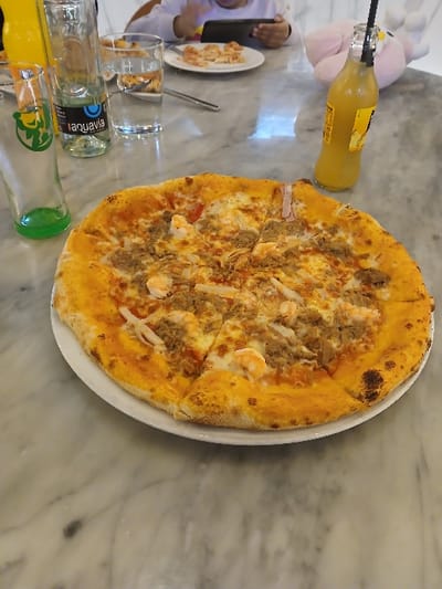 A whole pizza with tuna and shrimp toppings on a white plate dominates the view. The pizza is cut into slices. Several drink bottles, including what appears to be juice and water, are arranged near the pizza. Part of a plate with food, a fork, and glasses are also visible. A pink stuffed animal sits in the background. The table has a marble-like surface.
