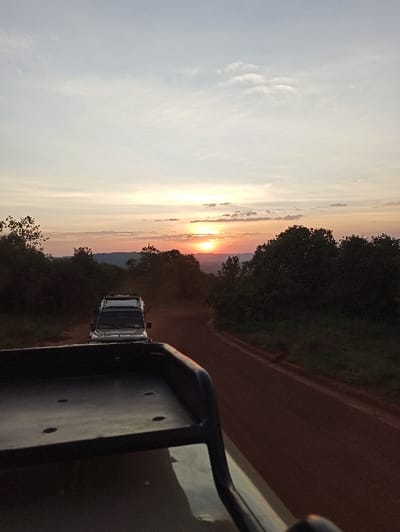 Captured from within a vehicle, the photo showcases a vibrant sunset. Orange and pink hues illuminate the horizon, partially obscured by silhouetted trees. Another vehicle trails behind, its presence adding depth to the landscape. The foreground displays part of the vehicle from which the photo was taken, providing a sense of perspective. The scene evokes a feeling of adventure and exploration.