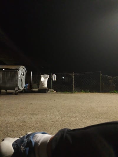 The image depicts an outdoor scene at night in Grohotno, Bulgaria. In the foreground, there is a partial view of a person wearing white socks, blue jeans, and a dark-colored jacket, suggesting they are lying down. Beyond them, there is an expanse of asphalt ground. In the middle ground, a large, gray dumpster with visible cans inside, a white bag on a platform, and a plastic bottle hung on a wire fence are visible. Green grass is seen growing at the base of the fence. The background is dark, suggesting it is nighttime. The scene appears to be in a public or semi-public space.