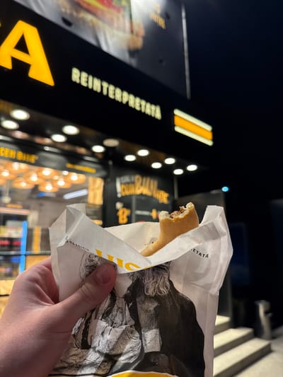 A first-person, low-light photograph depicts a human hand holding a partially consumed pastry within a white paper bag, positioned in front of a commercial establishment at night. The hand, with visible skin tone, grasps the lower-left section of the bag. The pastry, light brown and elongated, shows a distinct bite taken from one end, revealing a dark brown filling. The paper bag features yellow branding and a faded black-and-white graphic of a figure with long hair or a beard, with the word "TRADITIE" (Tradition) partially visible.

In the blurred background, the facade of a business is illuminated. A prominent black sign features the word "REINTERPRETATA" (Reinterpreted) in white, uppercase, illuminated letters, alongside a large yellow "A" to the left. Above this, a large, out-of-focus image of a food item, possibly a sandwich, is visible. Below the main sign, additional text "SEN BINE" and "DIN 1990" can be discerned on separate panels. The interior of the establishment shows warm, circular lighting fixtures and display cases containing various items, likely baked goods. To the right of the frame, a set of concrete steps leads upwards, and a cylindrical metallic object, possibly a trash receptacle, stands near the steps. The scene takes place at night in Agigea, Romania, at a "Reinterpretata" branded establishment, likely a bakery or fast-food outlet.