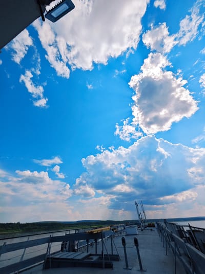 This media file captures a low-angle shot of a bright blue sky filled with fluffy white clouds, some of which are lit by sunbeams breaking through. In the foreground, the deck of a vessel is visible, with metal railings and various equipment such as poles and what appears to be a white utility box. The deck is constructed of concrete. In the distance, a tree-lined shore stretches out along a body of water, with rolling hills forming the horizon under the expansive sky.
The scene suggests a daytime setting, likely late morning or afternoon, given the bright sunlight and clear sky. The weather appears fair with some scattered clouds. The location cues, particularly the river and the dense forest on the distant banks, combined with the provided context of Podkamennaya Tunguska, Russia, point to a natural, possibly remote environment. There are no people visible in the media. The overall mood is serene and expansive, emphasizing the vastness of the sky and the natural landscape. There is no visible text in the media file.