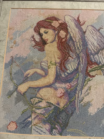 The image shows a framed cross-stitch tapestry depicting a woman with wings, holding a bow and arrow. She has long, reddish-brown hair adorned with pink flowers. Her attire consists of a purple and blue draped garment. The background is a soft blend of pink, blue, and gray hues, with subtle hints of green foliage and pink flowers. The tapestry is framed by a thin, light-colored wooden border. The location of this tapestry within the city of Zmiiv, Ukraine, is not discernible from the image.