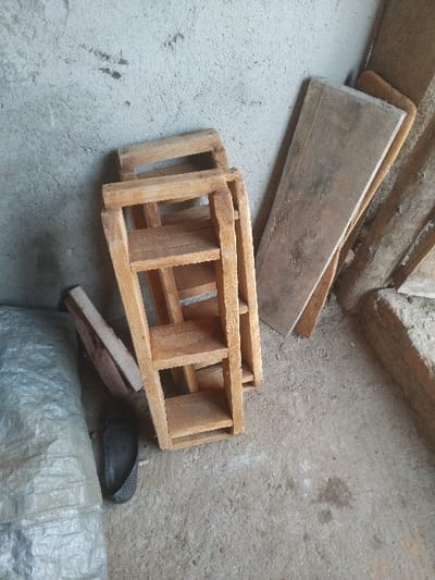 The image shows a cluttered corner. A weathered plank of wood rests against the wall. Below it, a small, open wooden frame with multiple sections sits askew. A smaller wooden piece is near a dark, perforated object. A large, silver tarp or plastic sheet fills the bottom right corner of the frame. The background is rough, concrete-like.