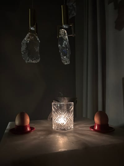 The image displays an indoor scene in the city of Баня, Bulgaria, illuminated by multiple artificial light sources. Two brass-toned overhead fixtures, each fitted with a downward-facing light, suspend large, irregularly shaped clear glass or crystal elements. Below, on a light-colored surface, possibly polished stone or marble, a central cut-glass tumbler with a diamond pattern emits light, casting radial patterns onto the tabletop. To the left of this central light source, a brown egg rests in a round, red egg cup with a wide brim. An identical brown egg in an identical red egg cup is positioned to the right. In the background, behind the central tumbler, a metallic bucket or ice pail is partially visible, containing a utensil, possibly tongs. The background consists of a dark wall area on the left and vertically pleated drapes on the right. No people are present.