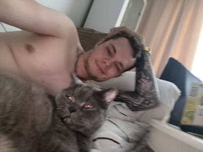 A man and a cat are seen in a close-up, slightly blurry shot. The man, with tattoos on his arm and a gold chain around his neck, is looking towards the camera with a slight smile. His face is partially obscured by the cat. The cat, a grey feline with amber eyes, is resting on the man, its head close to his face. The background suggests an indoor setting, possibly a bedroom, with curtains and what appears to be a shelf with some items on it. The image is oriented sideways, making it difficult to discern the full scene. No specific time of day or weather conditions are evident. The overall mood is intimate and relaxed.