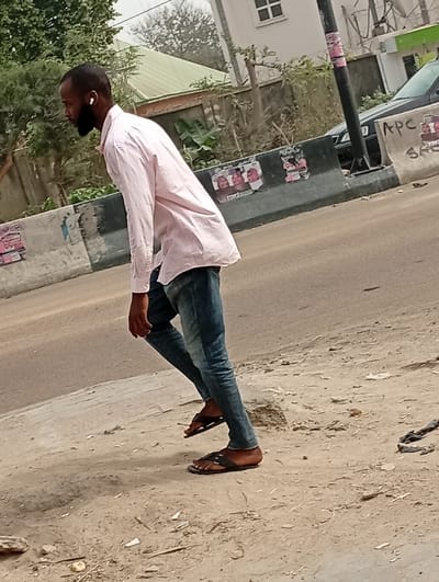 An adult male is depicted walking on an unpaved, sandy-dirt surface in the city of Darmanawa, Nigeria. He possesses dark skin, a short dark haircut, and a black beard, and is wearing a light pink long-sleeved button-up shirt, blue denim jeans, and black sandals. A white wireless earbud is visible in his right ear. He is in profile, stepping forward with his left foot planted and his right foot elevated. To the immediate right and behind the subject is an asphalt road, separated by concrete barriers. These barriers bear multiple posters and "APC" graffiti. Behind the barriers, a partially visible black vehicle is present, along with a black utility pole and dense green foliage, including trees and broad-leaf plants. Further in the background, a building with a light green sloped roof and a white wall featuring a rectangular window is visible.