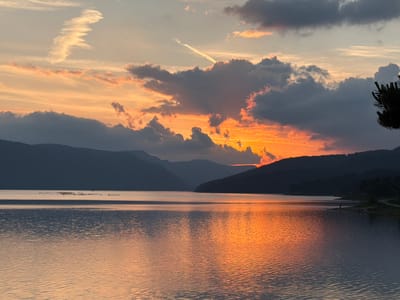 This image depicts a sunset over Lake Dospat in Dospat, Bulgaria. The sky is dominated by a mix of warm orange, red, and pink hues near the horizon, transitioning to lighter blues and grays at the top. Dark, voluminous cumulus clouds are prominent, heavily backlit by the setting sun, creating bright orange and red edges. Two distinct contrails are visible: one wavy, elongated contrail in the upper left, and another straight, diagonal contrail in the upper center-right.

Below the sky, multiple layers of dark, silhouetted mountains or hills stretch across the entire horizon, defining the far edge of the lake. The lake's surface reflects the intense orange and red colors of the sunset, particularly in the central and right portions, with gentle ripples disturbing the reflections. In the mid-ground on the left, a cluster of small, dark objects, possibly birds, rests on the water.

On the far right, a dark silhouette of coniferous tree branches extends into the frame from the upper right corner. The near shoreline is visible on the right, where a single human figure stands facing the water, partially silhouetted against the bright reflection. A paved path or road runs along the shoreline, curving towards a forested area in the distance. The overall scene is characterized by the dramatic contrast between the brilliant sunset colors and the dark silhouettes of the landscape.