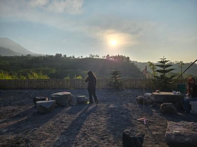 In Cangkringan, Indonesia, at sunrise, two women are present in an outdoor seating area. One woman, wearing a dark maroon top and dark pants, stands in the center of the frame, facing away from the camera, with her back to a small pine tree. The second woman, wearing a brown hijab and a dark maroon top with blue jeans, sits on a bench to the right of the frame, looking to her left. The ground is covered in dark, sandy soil. Several large rocks are scattered around the area, some arranged as seating around a low stone table. A wooden fence borders the seating area, behind which is a hillside covered in lush green vegetation and trees. The sun, low in the sky, casts long shadows across the scene. The sky is a pale blue with scattered clouds.