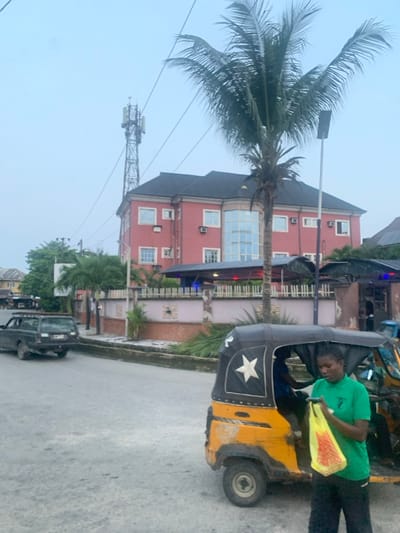 A three-wheeled vehicle, painted yellow and black with a white star, is parked on a street. A person in a green shirt stands nearby holding a yellow object. A palm tree towers over a multi-story pink building with numerous windows. A utility tower with wires extends into the sky. A black pickup truck sits on the street, and another building is visible in the background. The scene appears to be a street view.