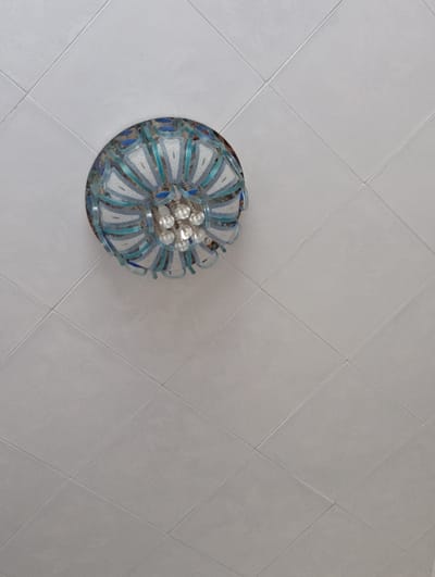 A close-up, high-angle shot shows a decorative ceiling fixture with a swirling, fan-like design. The fixture has multiple layers of curved, translucent material in shades of white and blue, interspersed with small, clear crystalline elements that appear to be glass or crystal. The ceiling itself is tiled with square, light gray tiles arranged diagonally, creating a grid pattern. The overall impression is of an interior space, likely a room with a decorative ceiling. The image does not contain any people or provide any contextual information regarding its specific location in Fayzabad, Tajikistan.